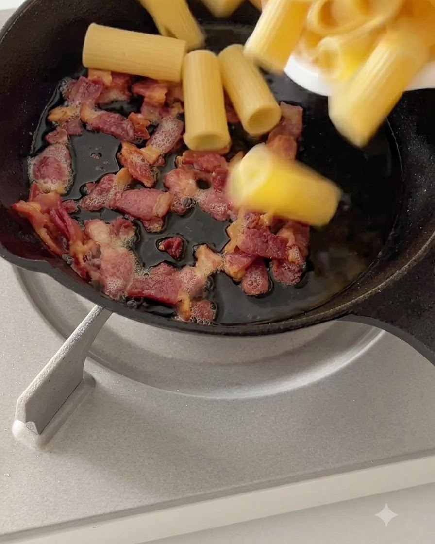 Gekochte Rigatoni-Pasta wird mit gekochtem Speck in einer Pfanne bei schwacher Hitze vermengt.