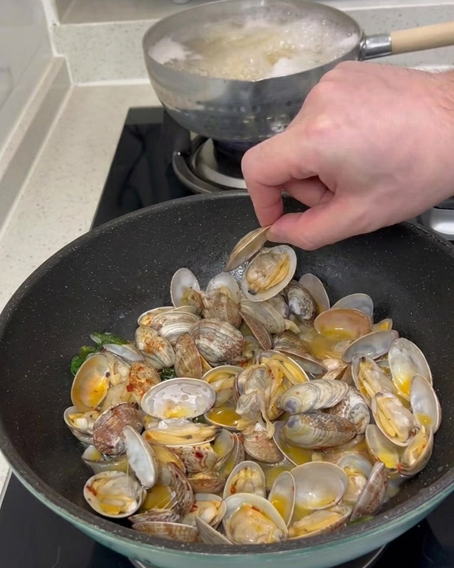 Die Hand einer Person hebt eine geöffnete Muschelschale aus einer Pfanne mit gekochten Muscheln und Soße.