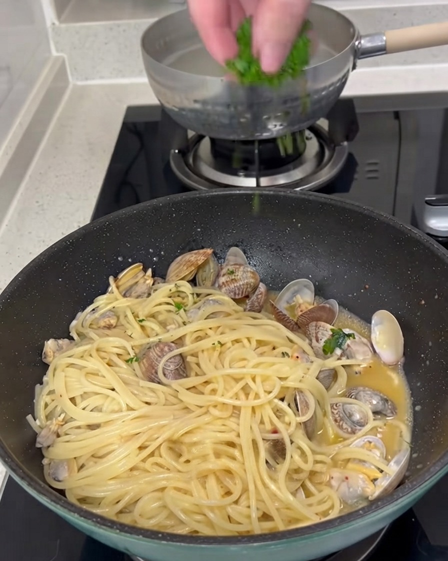 Frische gehackte grüne Petersilie wird von oben auf eine Pfanne mit gekochten Spaghetti und Muscheln gestreut.