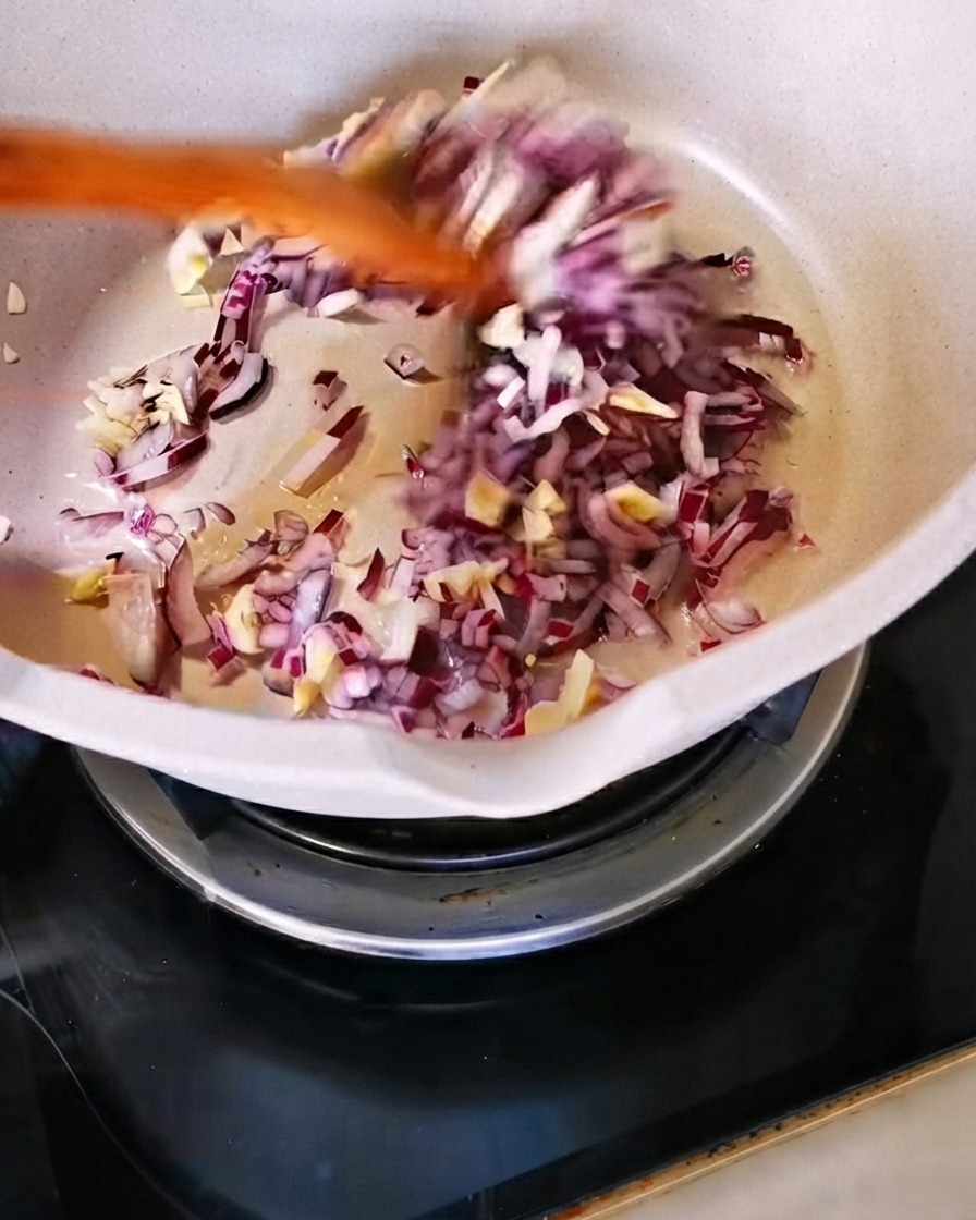 Gewürfelte rote Zwiebel und gehackter Knoblauch werden mit einem Holzspatel in einer weißen Pfanne angebraten.