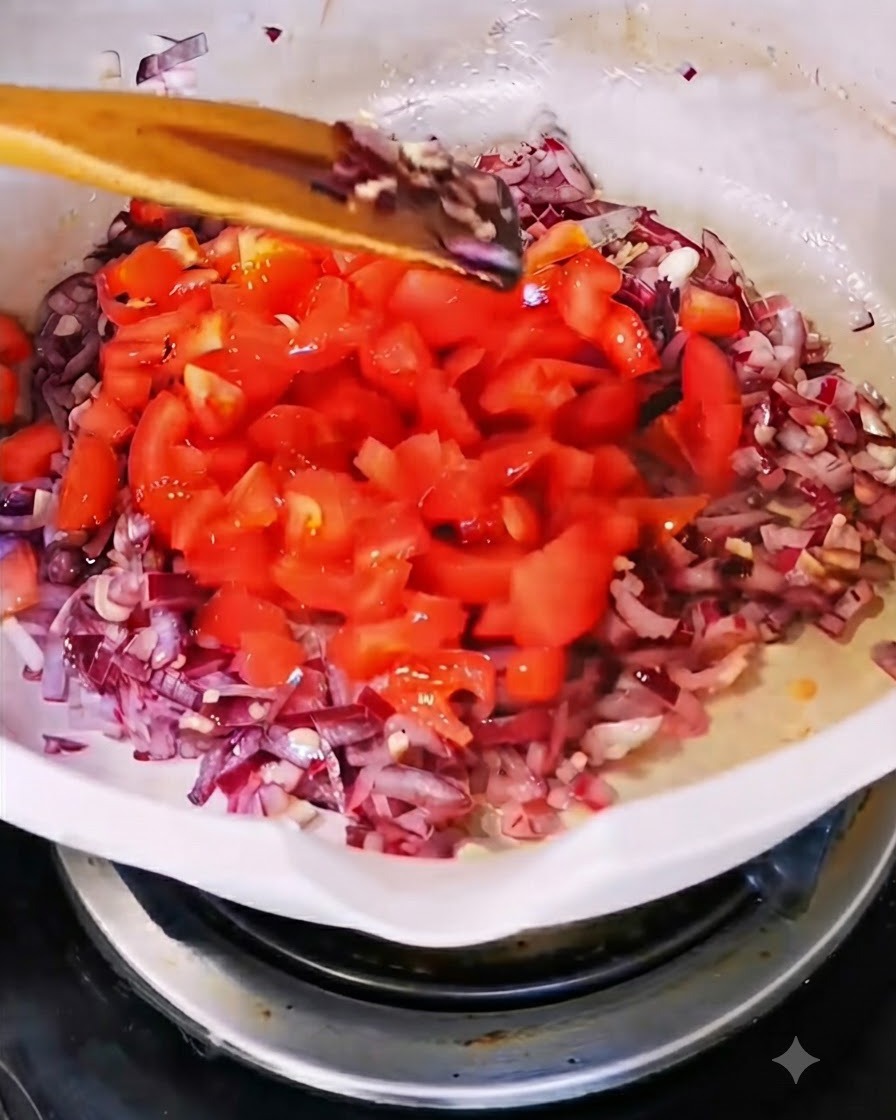 Frisch gewürfelte rote Tomaten werden in eine weiße Pfanne mit angebratenen Zwiebeln und Knoblauch gegeben.
