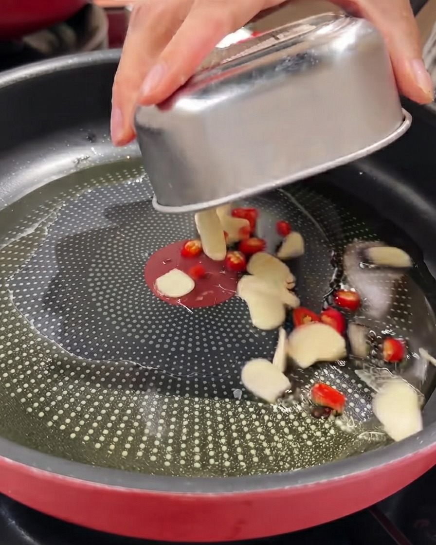 In Scheiben geschnittener Knoblauch, rote Vogelaugenchilis und Schnittlauch werden aus einem Metallbehälter in eine geölte Pfanne gegeben.