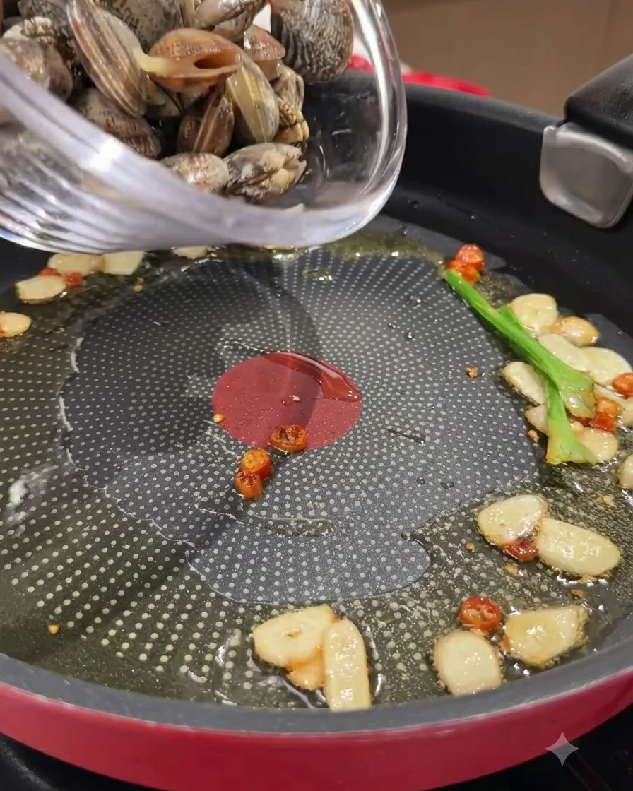 Frische Muscheln werden aus einer Glasschüssel in eine Bratpfanne mit sautiertem Knoblauch, Chilis und Öl geschüttet.