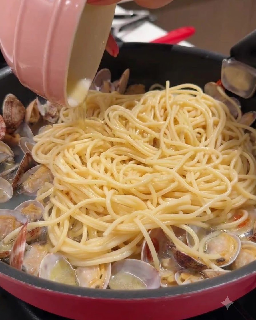 Eine kleine rosa Schüssel gießt Flüssigkeit in eine mit Spaghetti und Muscheln gefüllte Pfanne.