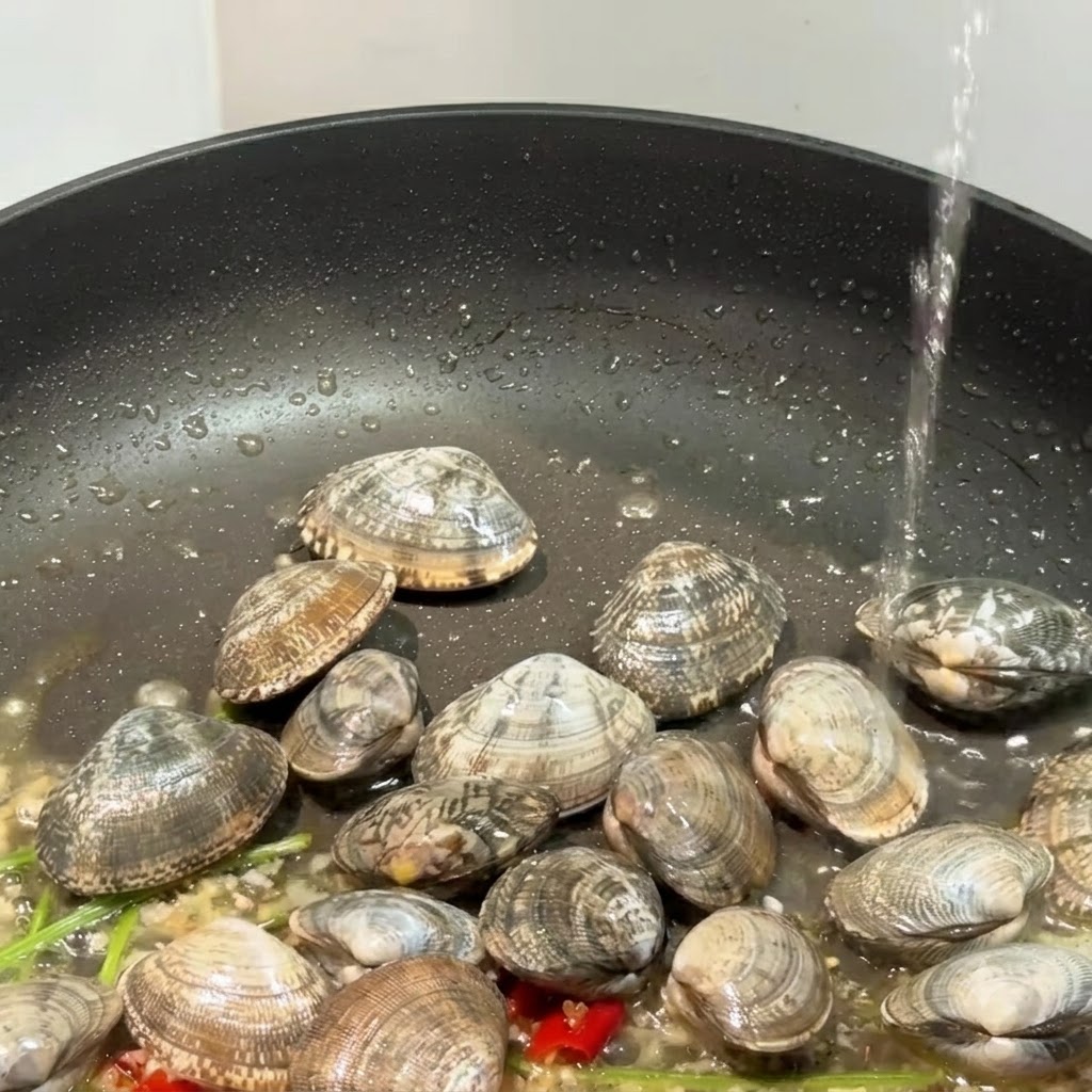 Eine schwarze Pfanne mit Muscheln, Knoblauch und Chilischoten, in die Flüssigkeit gegossen wird.
