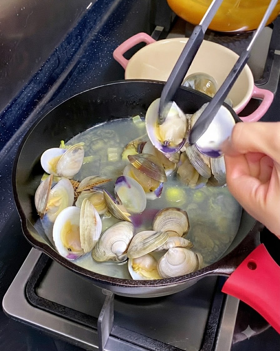 Eine Küchenzange hält eine geöffnete Muschelschale mit gekochtem Fleisch darin über einer Pfanne mit köchelnden Muscheln und Brühe.