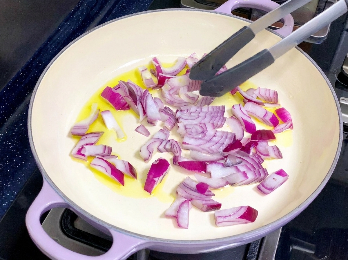 Eine helle Pfanne mit Olivenöl und gehackten roten Zwiebeln, die mit einer Zange umgerührt werden.