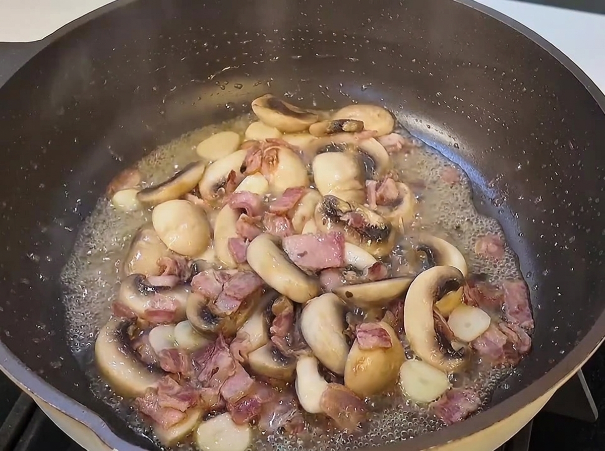 Gekochte Spaghettistränge werden in eine große Pfanne mit angebratenen Pilzscheiben und Speckstücken gegeben.