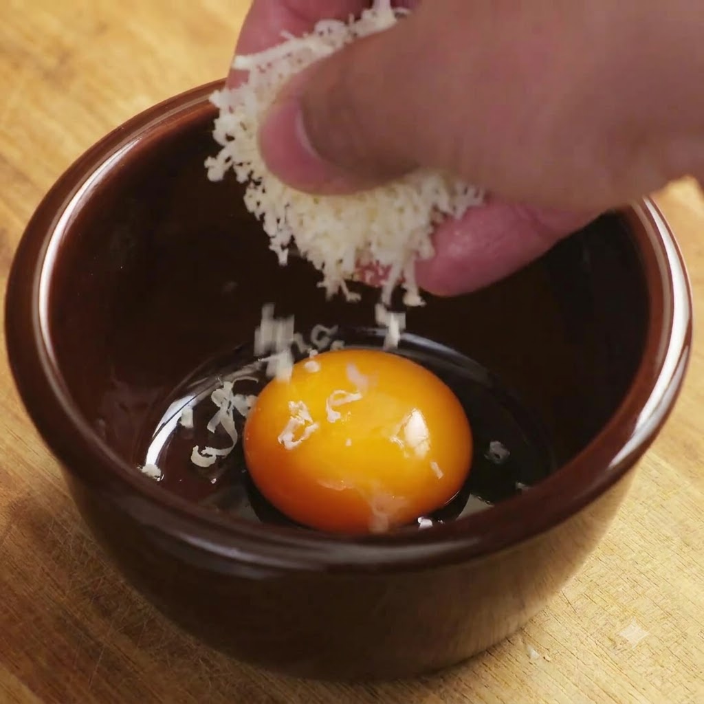 Geriebener Parmesan fällt auf ein helles Eigelb in einer braunen Keramikschüssel.