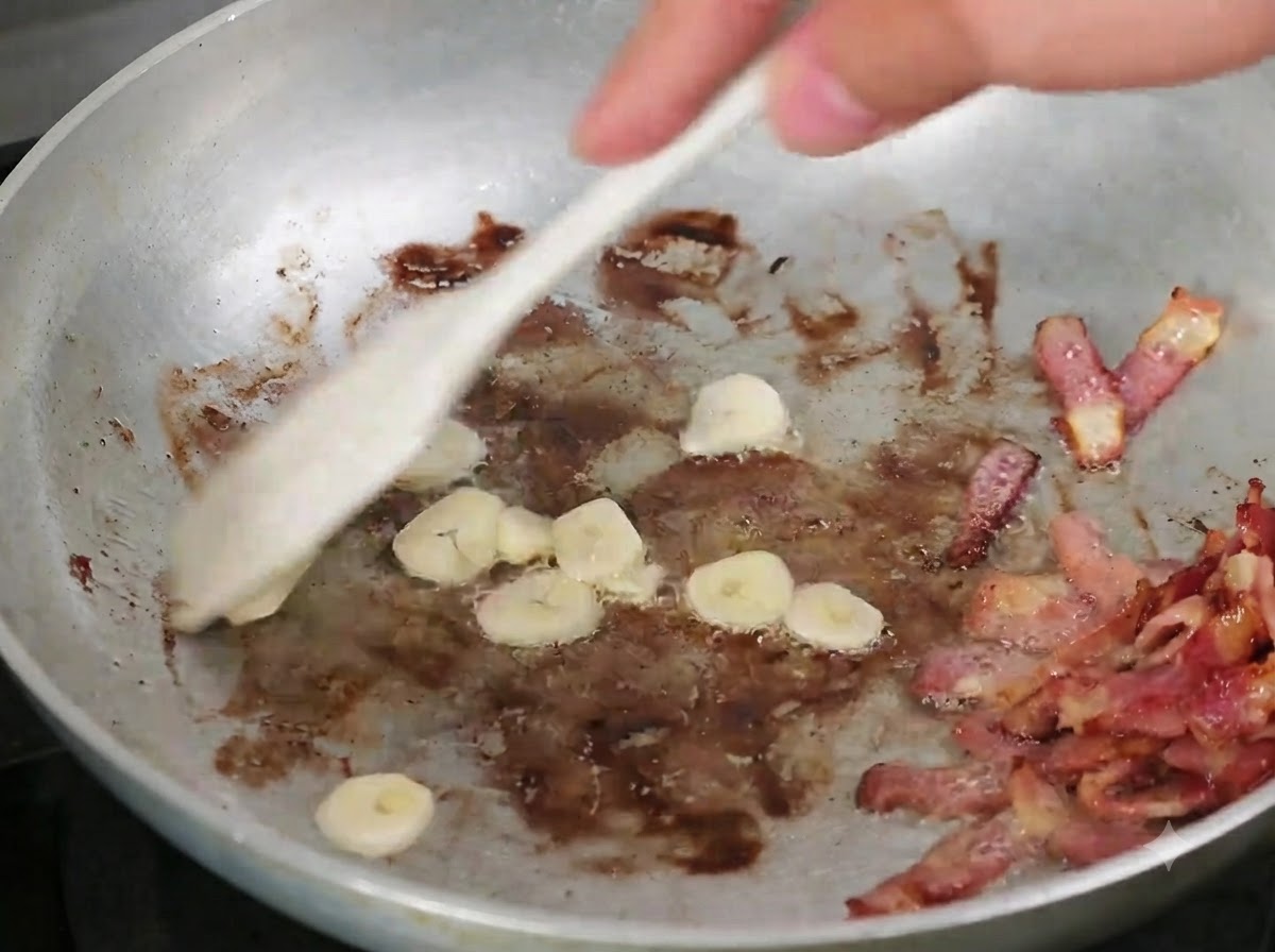 Knoblauchscheiben werden mit einem Holzlöffel unter den gebratenen Speck in der Pfanne gemischt.