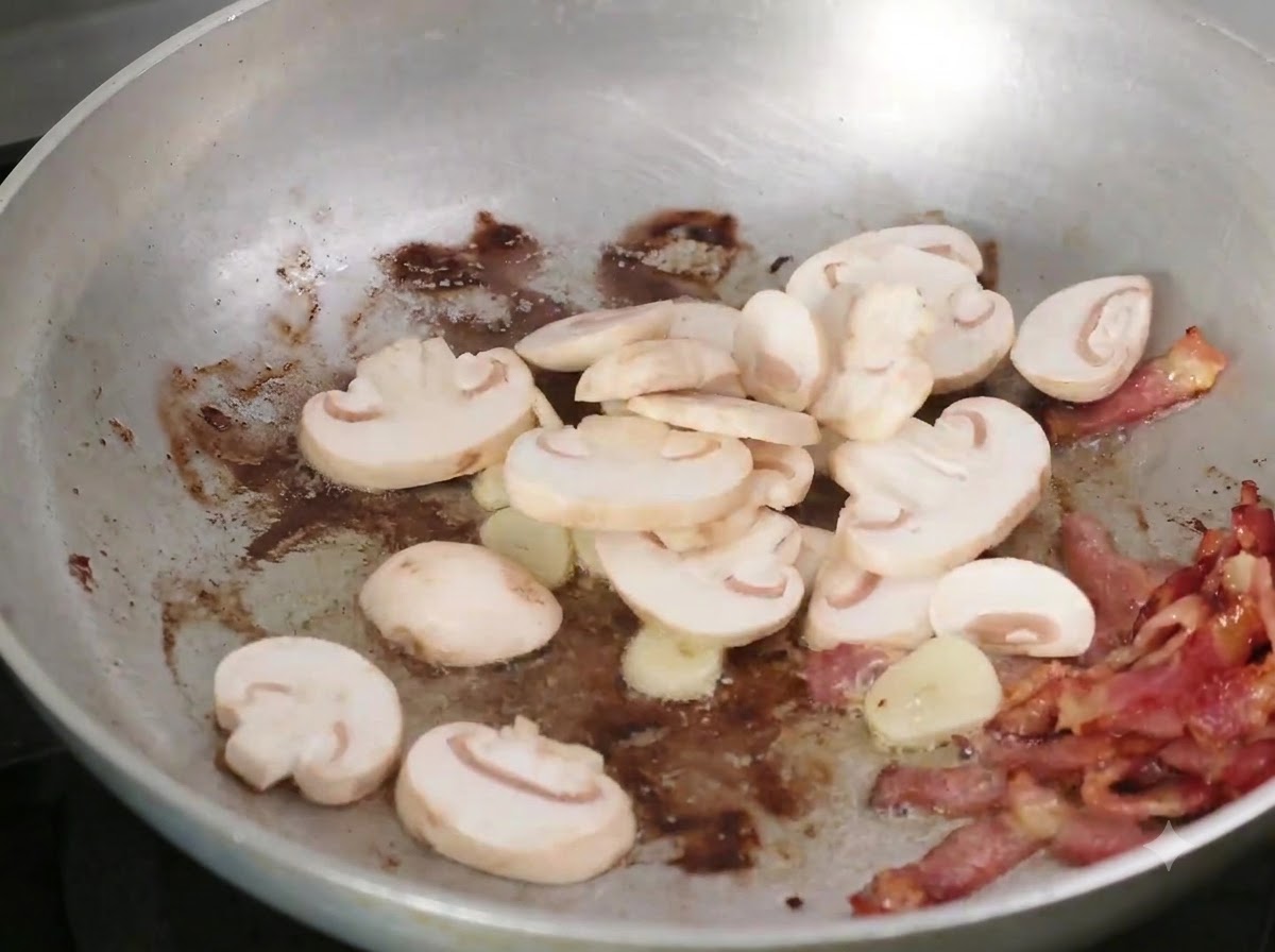 In Scheiben geschnittene Pilze werden zum Speck und Knoblauch in die Bratpfanne gegeben.