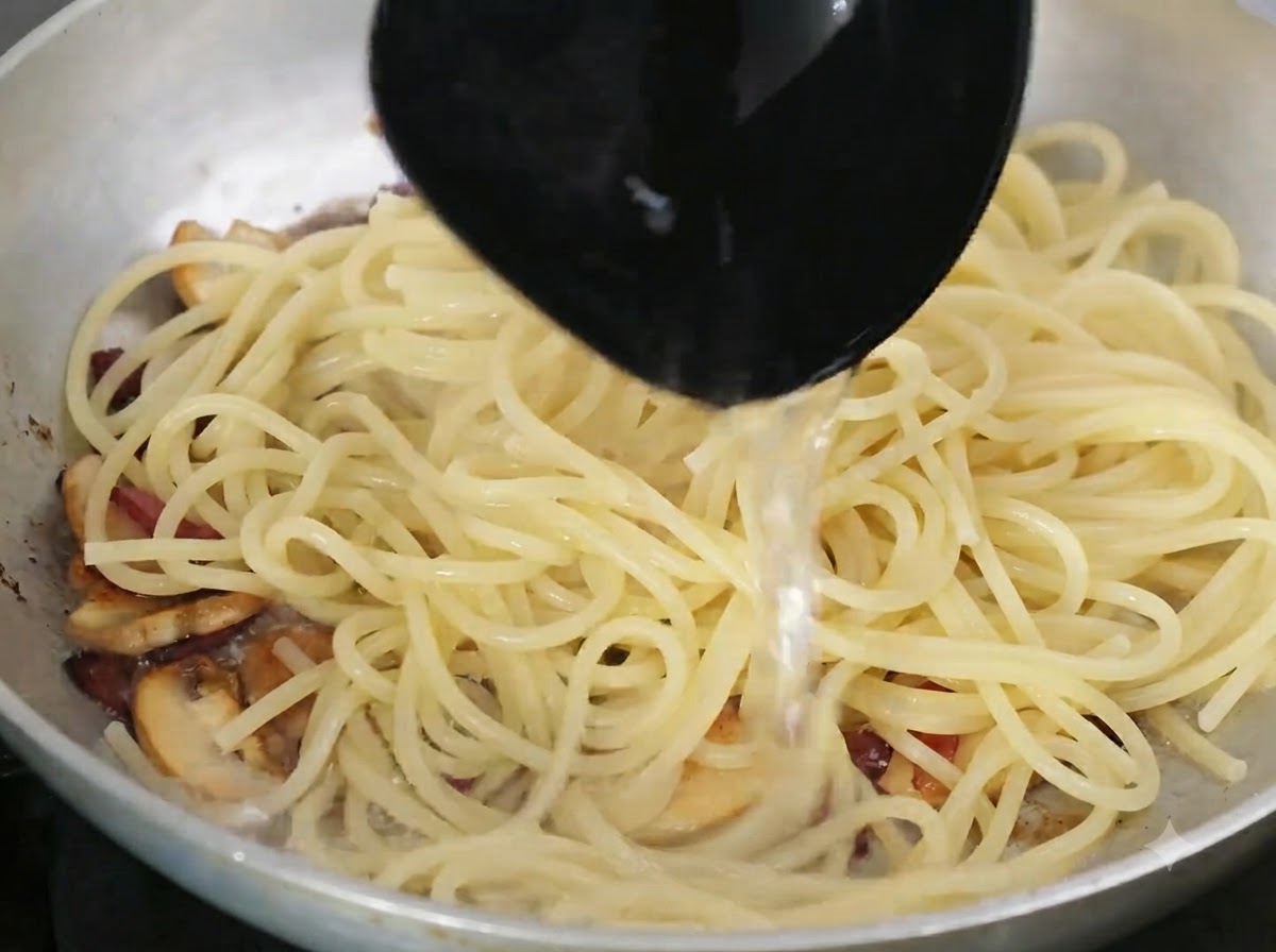 Eine Schöpfkelle gießt stärkehaltiges Nudelwasser in die Pfanne mit der Pasta und den Zutaten.