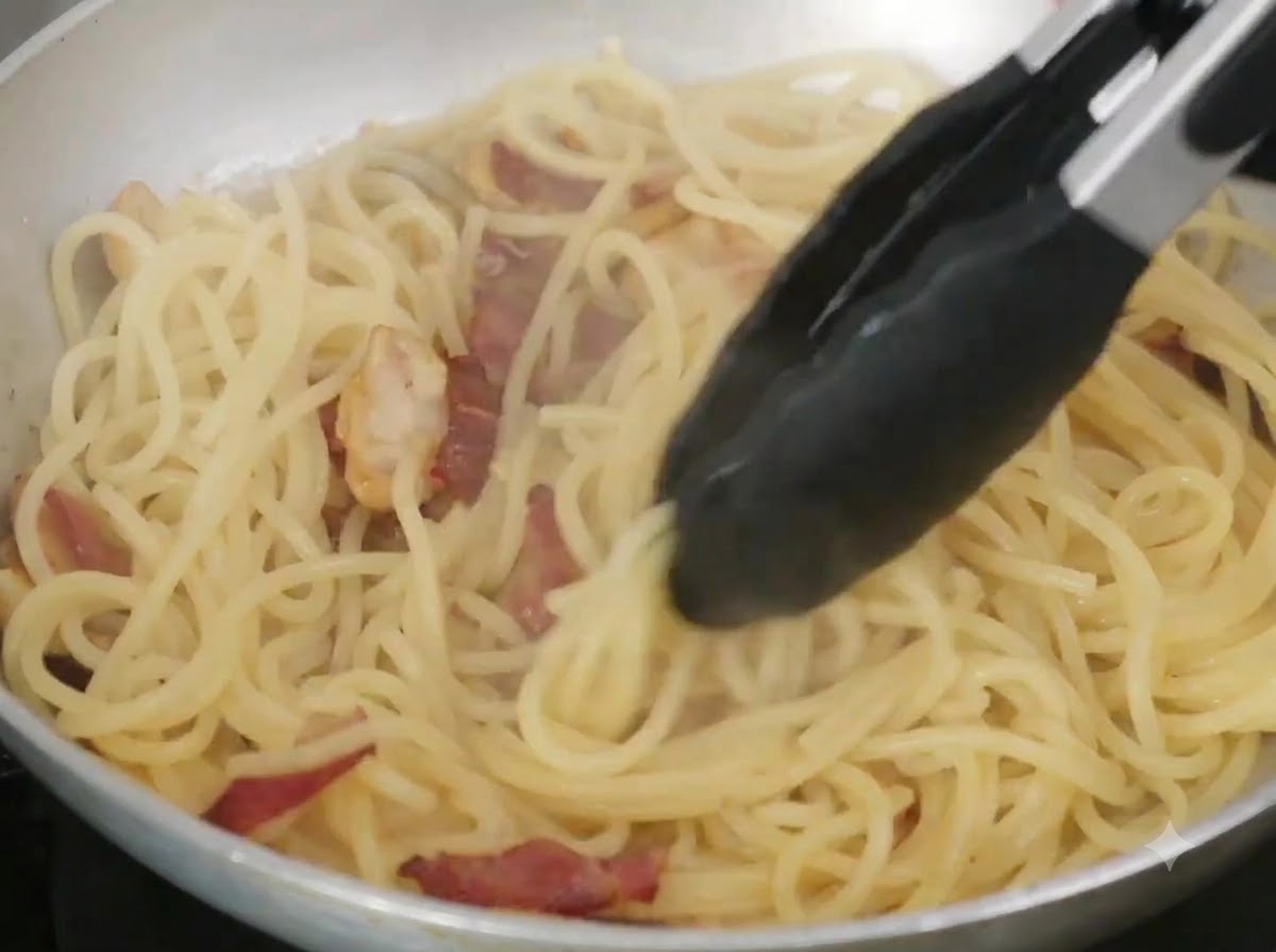 Butter und Nudelwasser werden mit einer Zange in einer Bratpfanne unter die Spaghetti gemischt.
