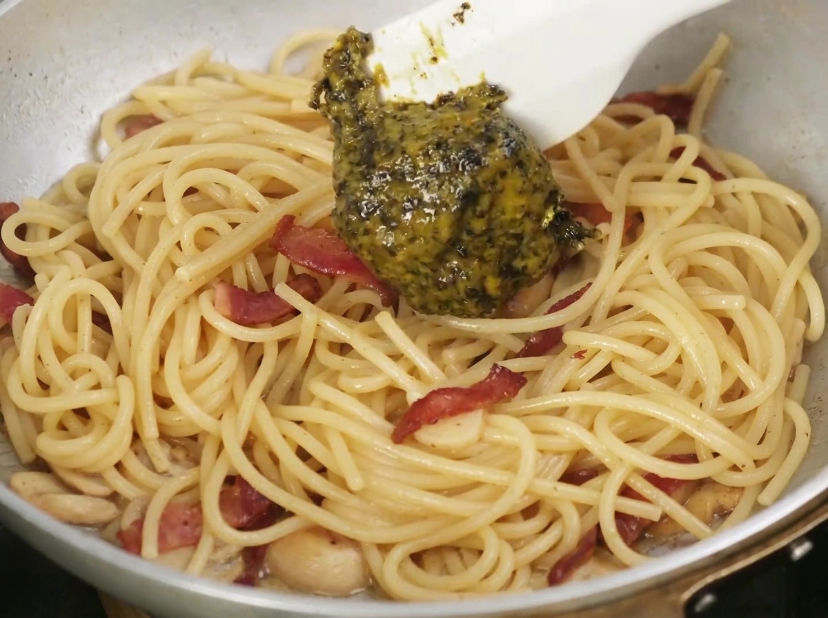 Schwarze Trüffelsoße wird in eine Pfanne mit gekochter Pasta und Speck gelöffelt.