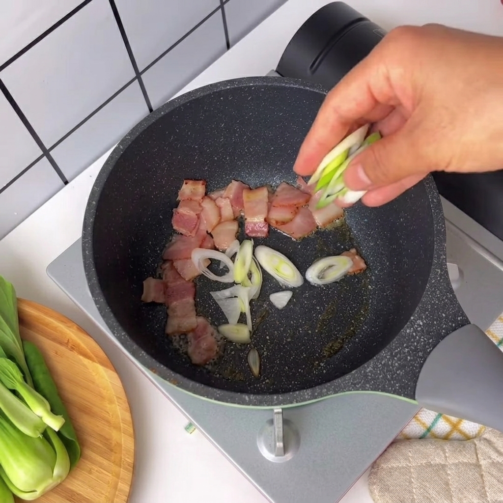 Gehackte Frühlingszwiebeln und Chilischote werden in eine Pfanne mit brutzelnden Speckstückchen gegeben.