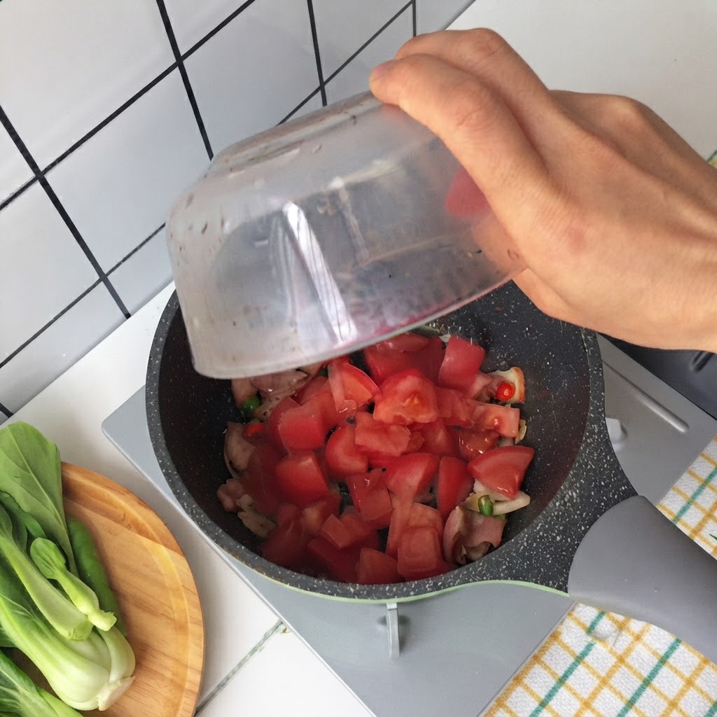 Hand gießt gehackte Tomaten aus einer Glasschüssel in eine Pfanne mit gebratenem Speck und Frühlingszwiebeln.