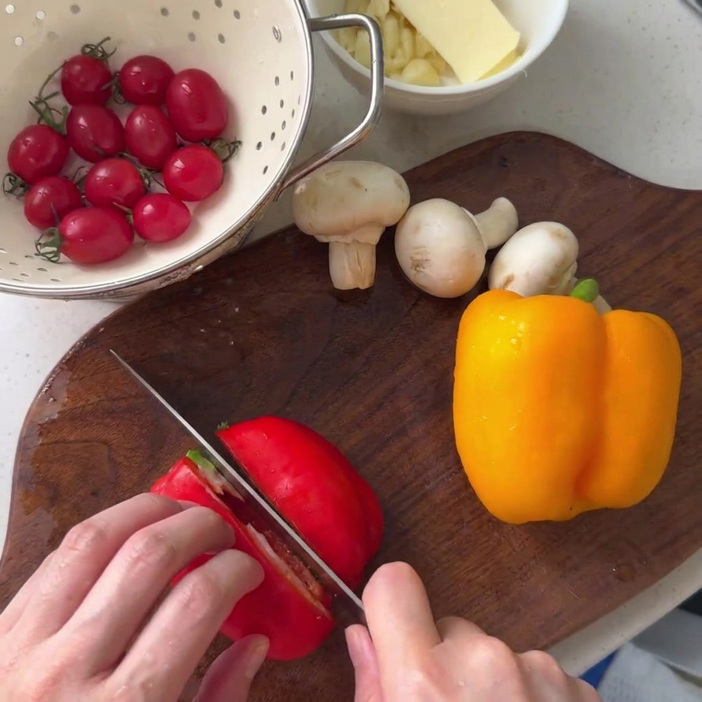 Rote Paprika wird auf einem hölzernen Schneidebrett neben einer gelben Paprika und Champignons geschnitten.