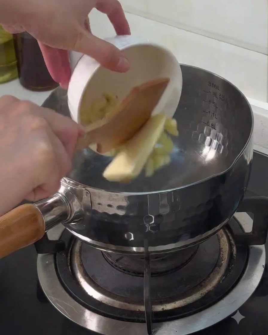 Gehackter Knoblauch und ein Stück Butter werden aus einer kleinen weißen Schüssel in einen Metalltopf auf dem Herd gegeben.