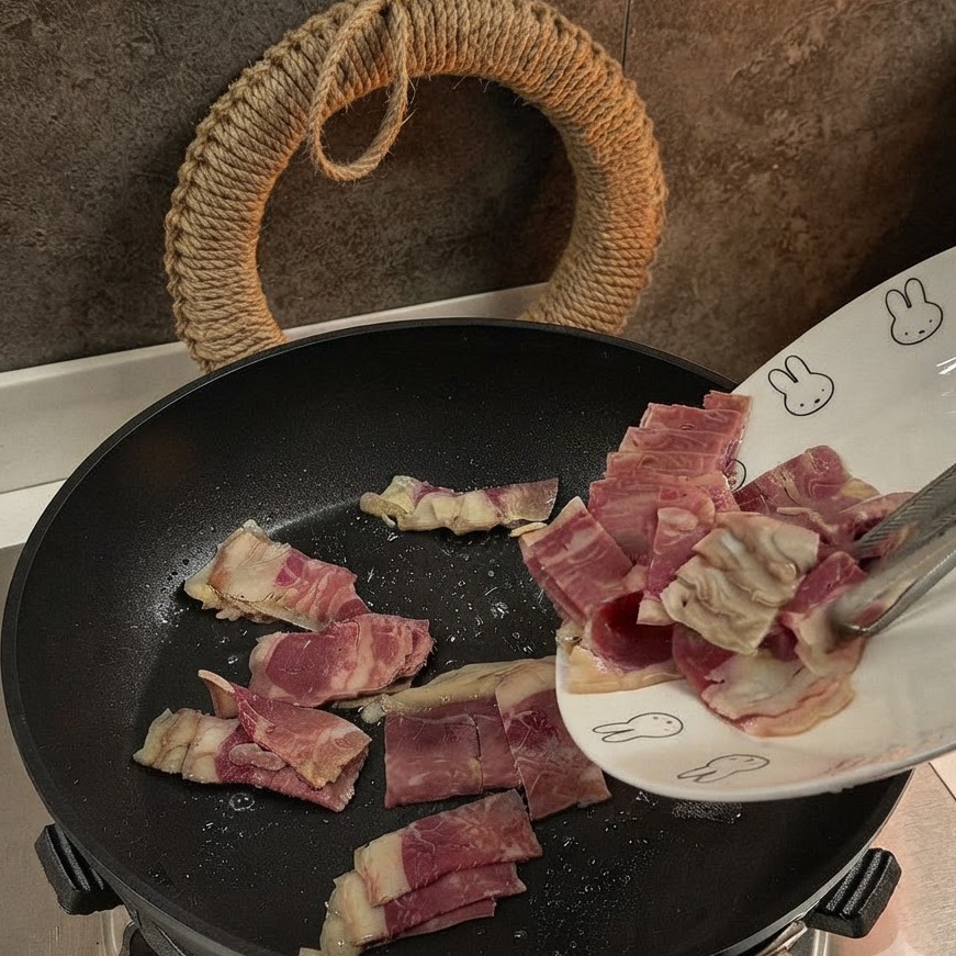 Geschnittene Speckstücke werden in eine schwarze Pfanne gegeben und darin angebraten.