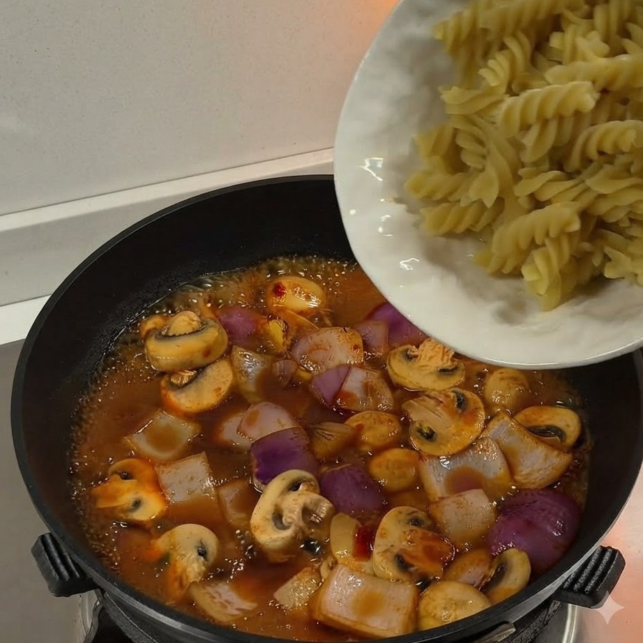 Gekochte Fusilli-Pasta wird von einem weißen Teller in eine Pfanne voller köchelnder Sauce und Gemüse geschüttet.