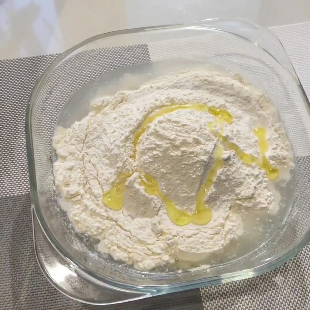 Olivenöl wird über Mehl und Wasser in einer Glasschüssel geträufelt.