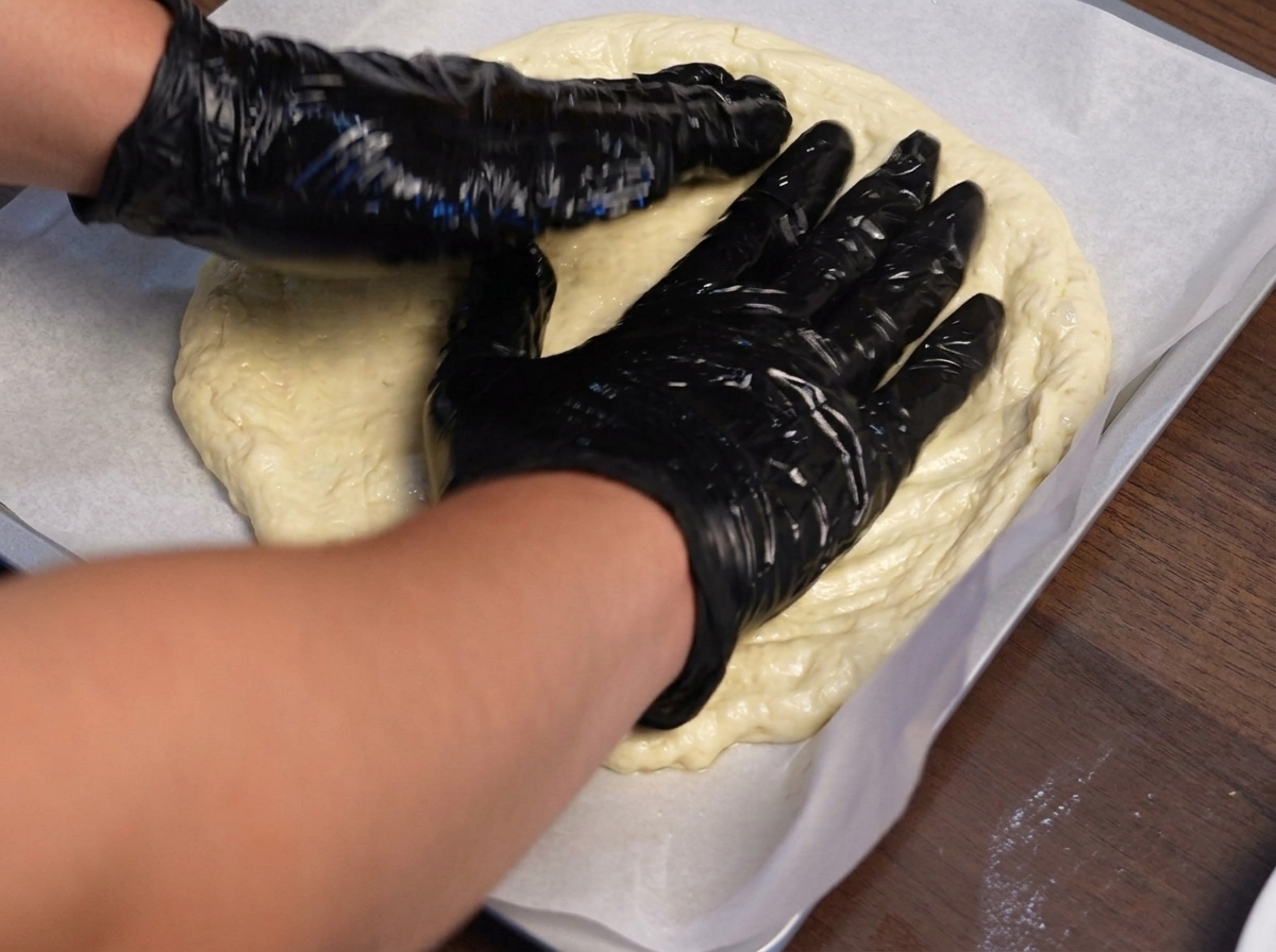 Hände mit Handschuhen formen Pizzateig auf einem mit Backpapier ausgelegten Backblech nach außen hin.