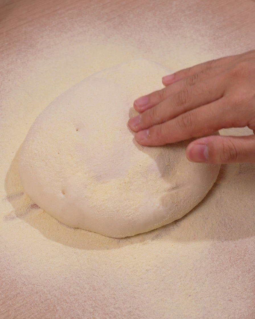 Eine Hand drückt sanft auf eine Teigkugel auf einer mit Maisgrieß bestreuten Arbeitsfläche.
