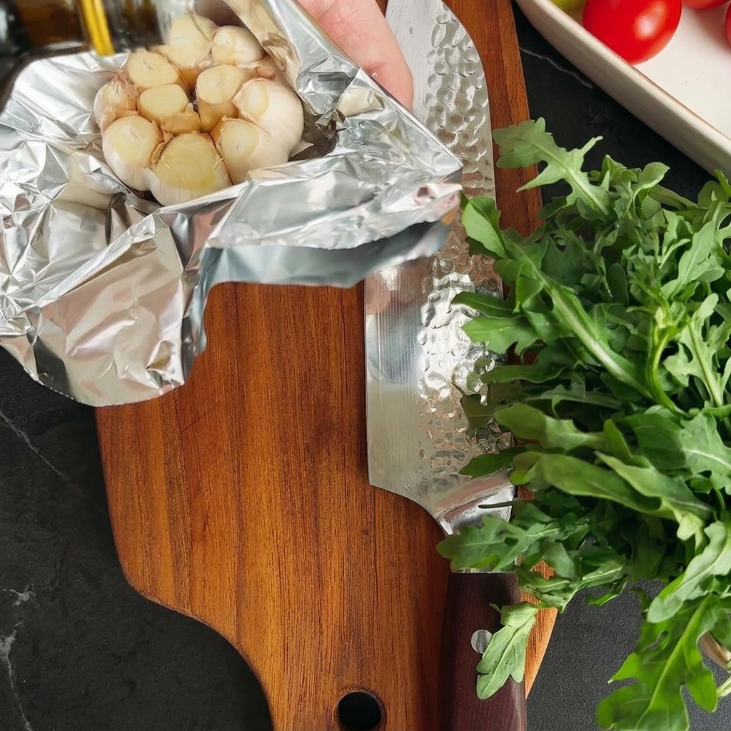 Olivenöl wird über eine unaufgewickelte Knoblauchknolle auf Aluminiumfolie geträufelt.