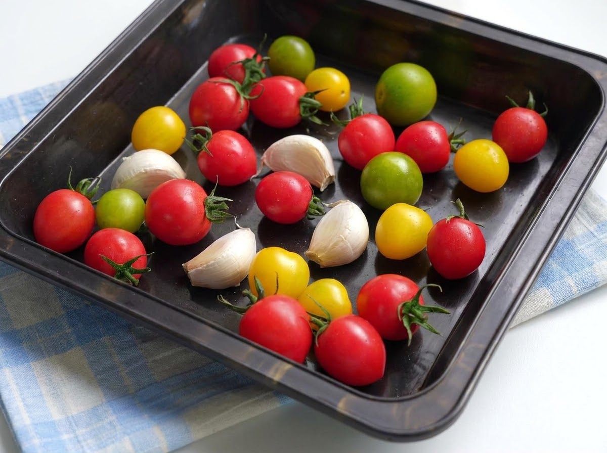 Ein Backblech gefüllt mit bunten Kirschtomaten und Knoblauchzehen, bedeckt mit Olivenöl und Gewürzen.