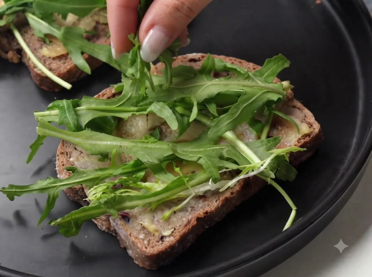 Eine Person schichtet Scheiben von gepökelter Schinken auf frischem grünem Rucola auf einer Brotscheibe.
