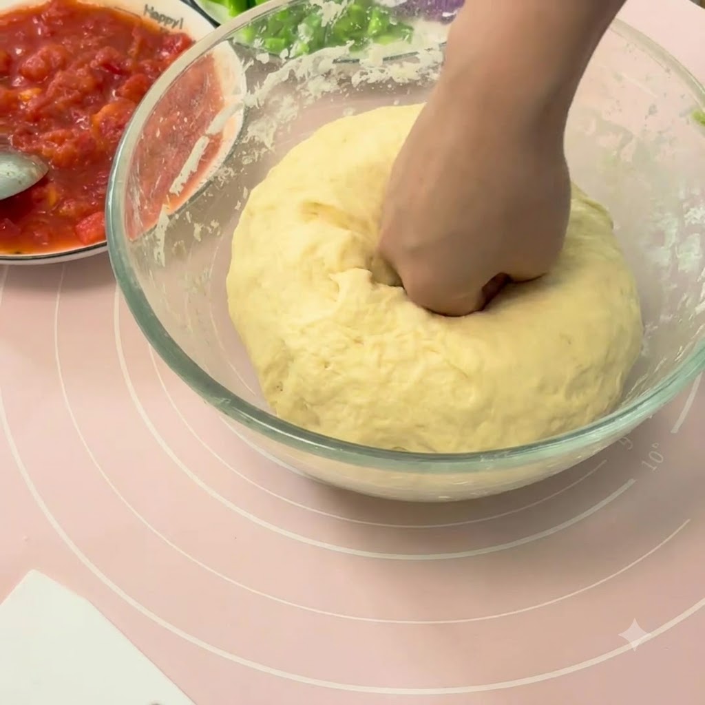 Eine Hand drückt eine große Kugel aufgegangenen Pizzateigs in einer Glasschüssel ein.