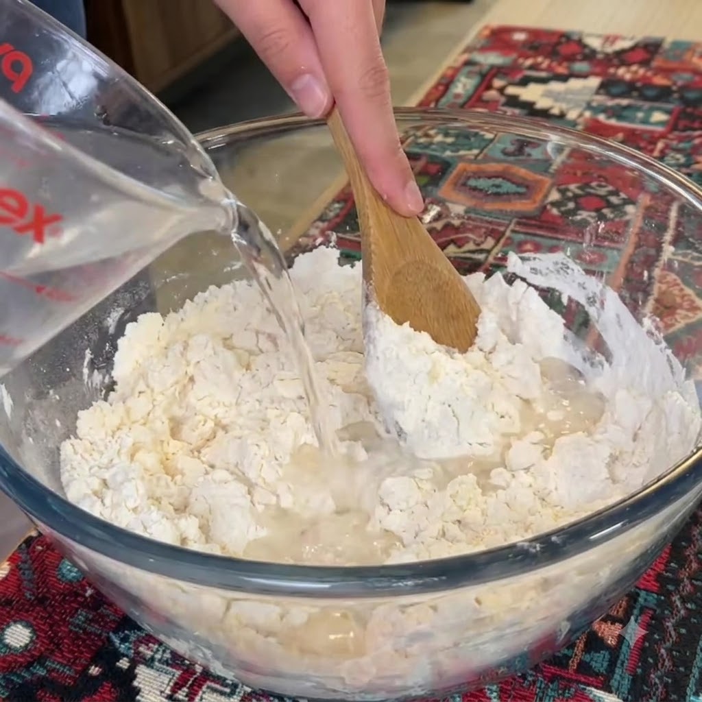 Wasser wird in eine Mehlschüssel gegossen, während ein Holzlöffel rührt.