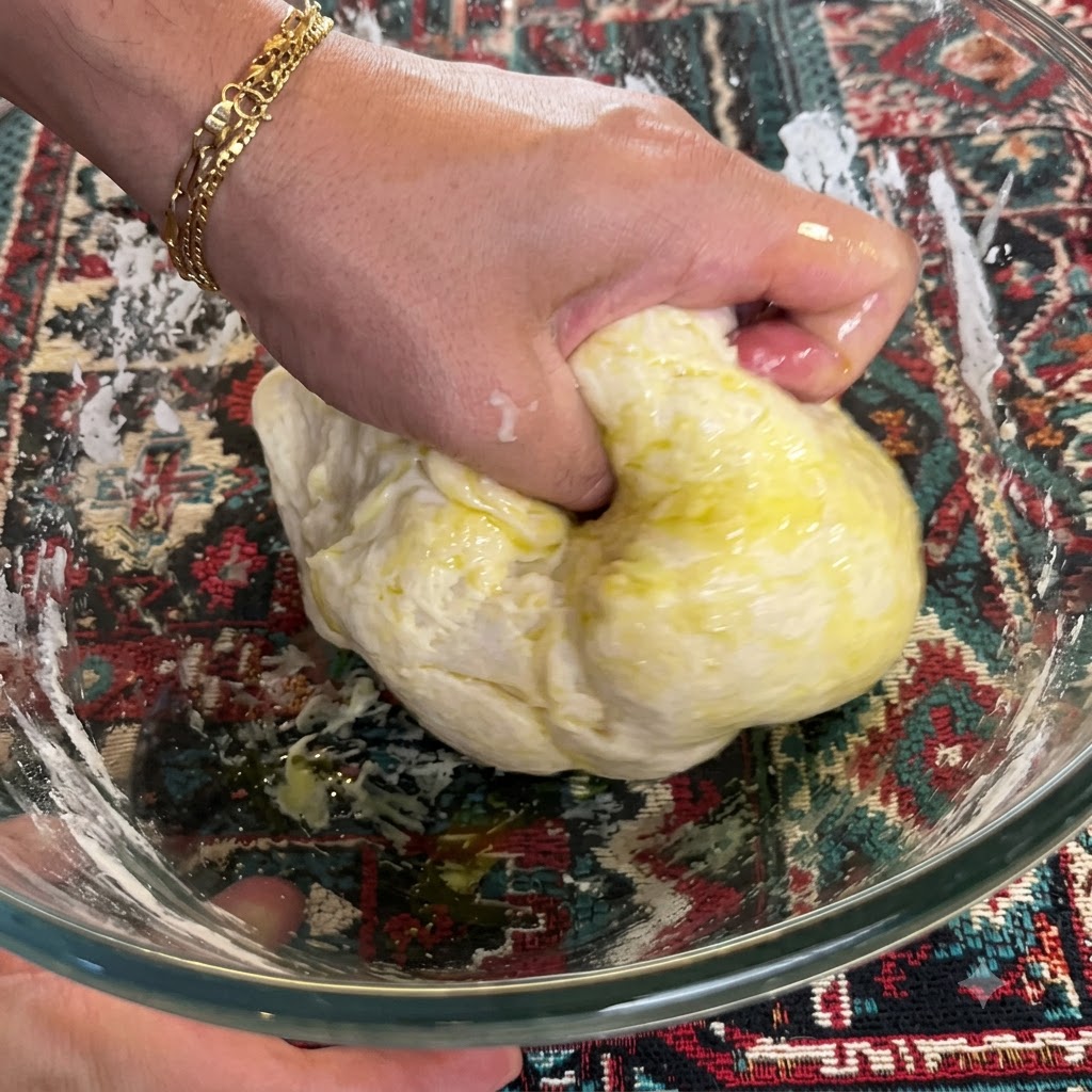 Eine Hand knetet und faltet Teig mit Olivenöl kräftig in einer Schüssel.