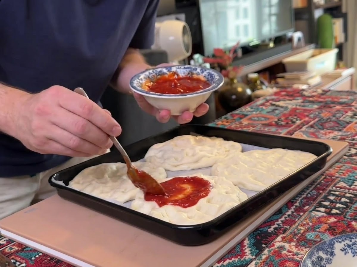 Ein Löffel verteilt rote Tomatensoße auf mehreren flachen Teiglingen auf einem Backblech.