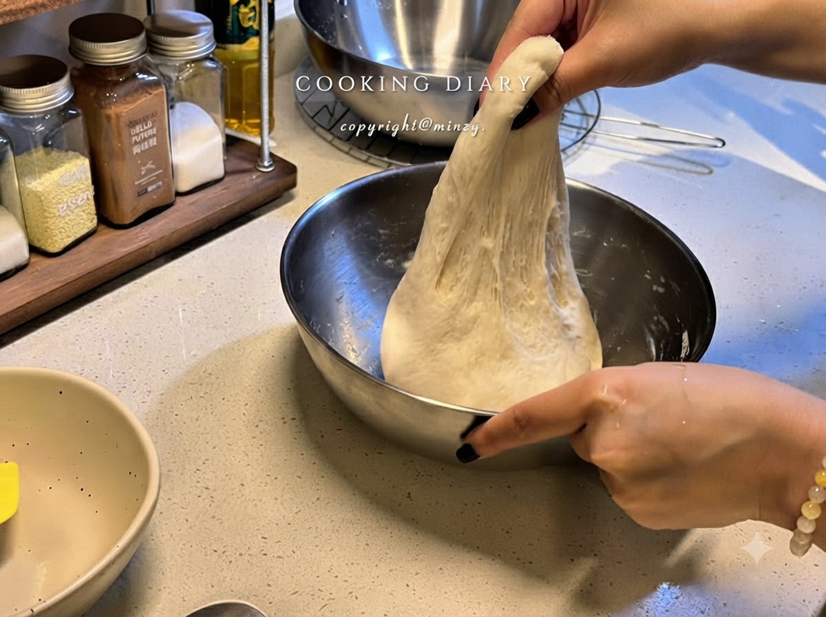 Zwei Hände heben eine Portion elastischen Pizzateigs in einer Metallschüssel nach oben, um ihn zu dehnen.