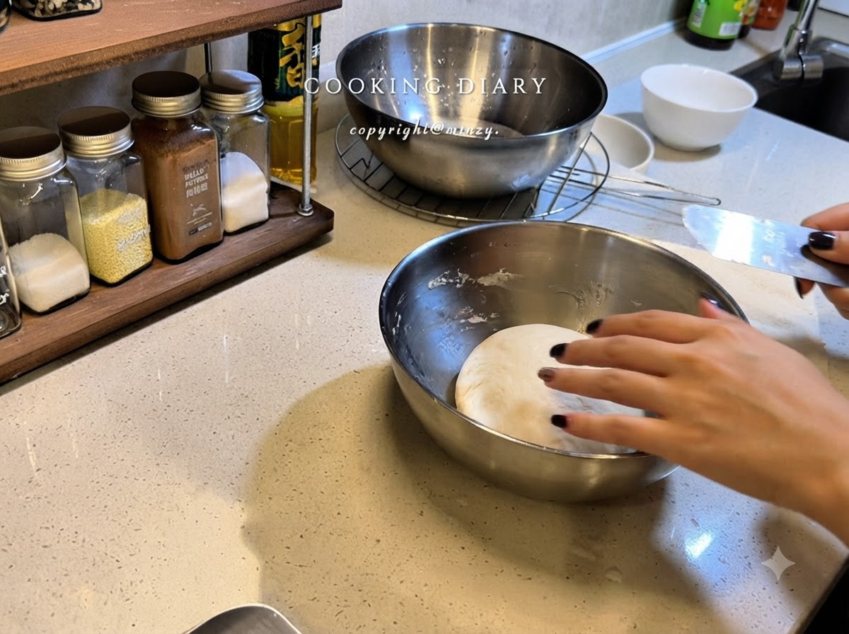 Eine Hand zieht Frischhaltefolie über eine Edelstahlschüssel, die eine runde Kugel Pizzateig enthält.