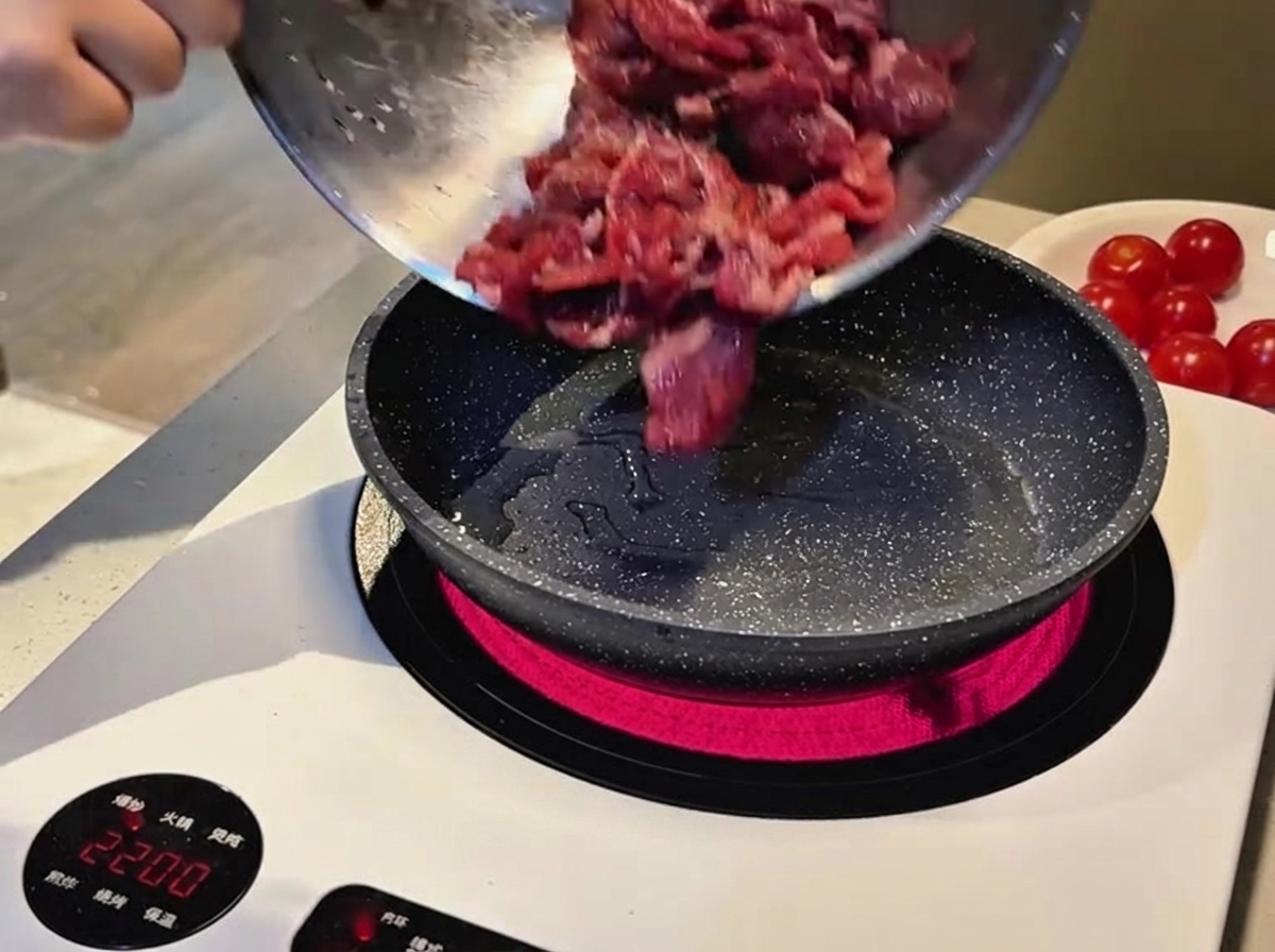 Rohes, mariniertes, in Scheiben geschnittenes Rindfleisch wird aus einer Metallschüssel in eine heiße gesprenkelte Bratpfanne auf einem Induktionsherd gegossen.