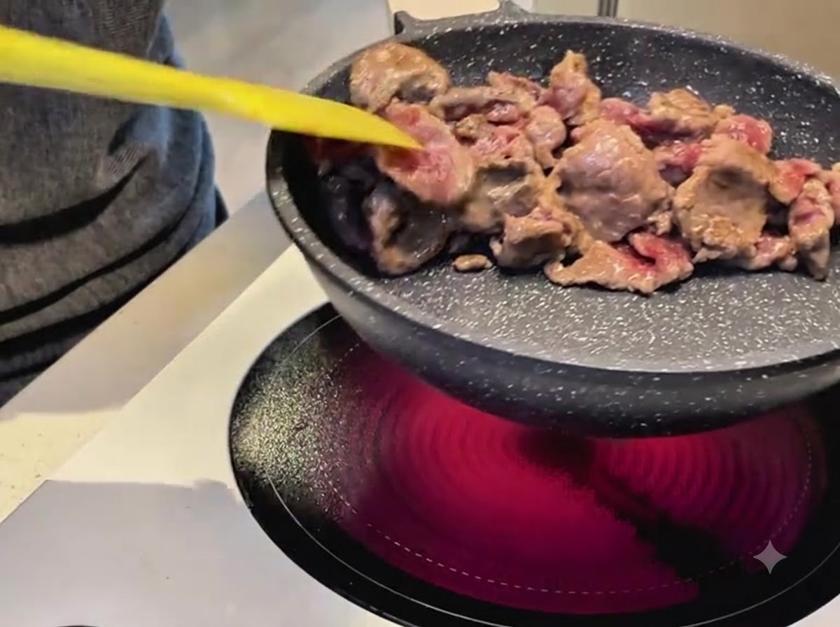 Mit einem gelben Spatel werden Rindfleischstreifen in einer beschichteten Pfanne auf einem Elektroherd gebraten.