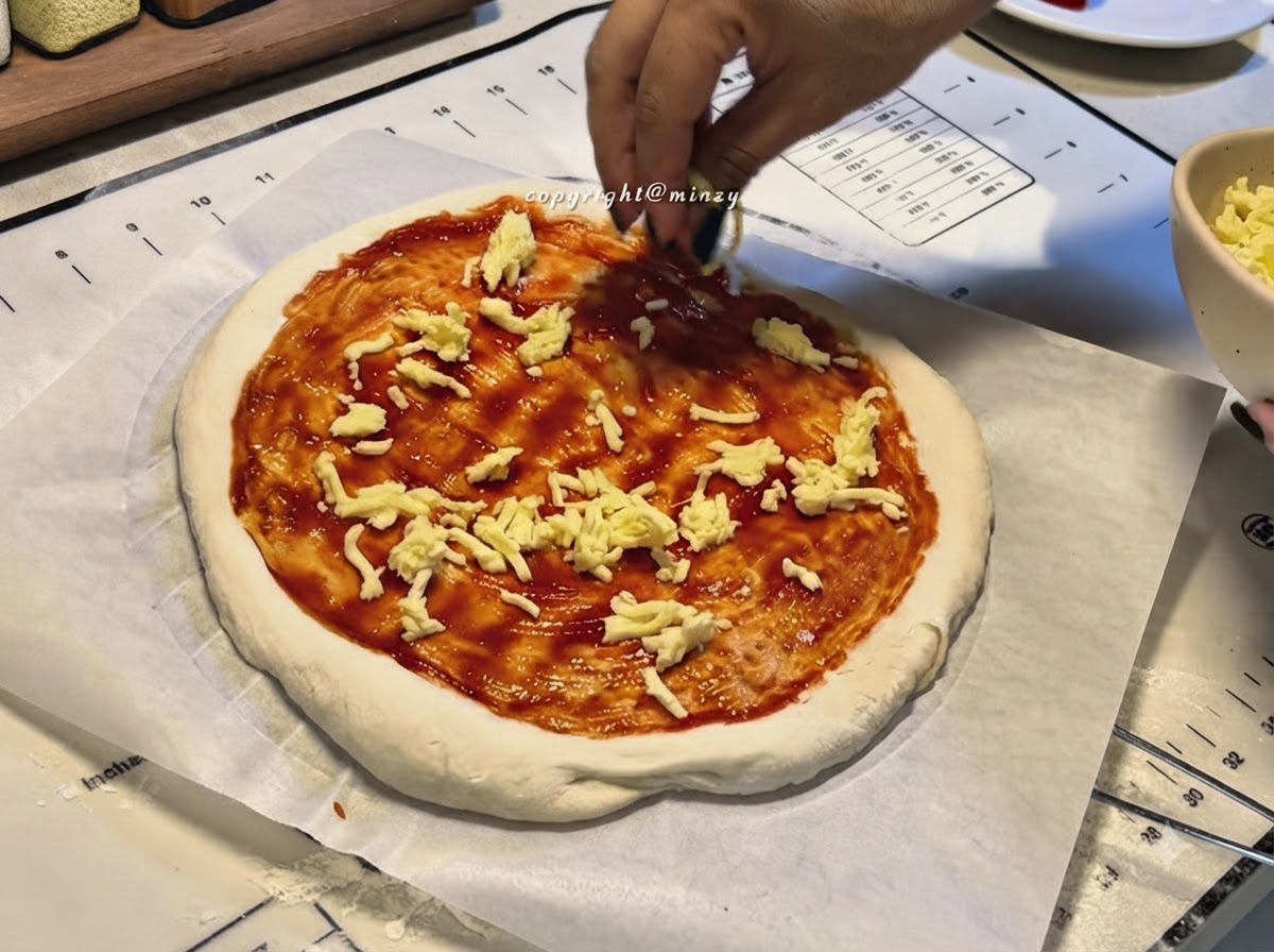 Hände streuen geriebenen Käse auf einen Pizzaboden, der mit roter Pizzasauce bestrichen ist.