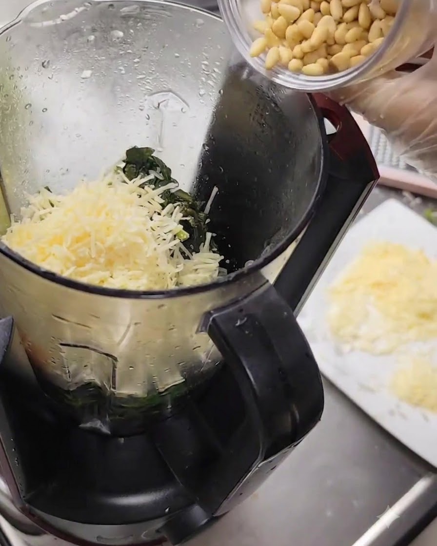 Pinienkerne werden aus einer kleinen Tasse in den mit Käse und Basilikum gefüllten Mixer geschüttet.