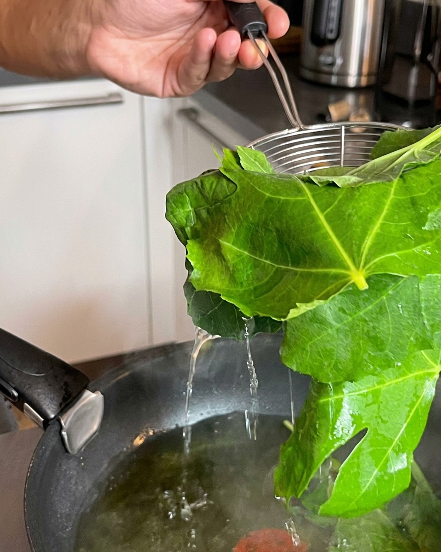 Ein Sieb hebt frisch blanchierte grüne Blätter aus einem kochenden Topf mit Wasser.