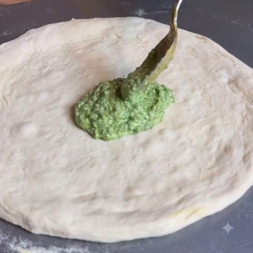 Ein Löffel verteilt grüne Pestosoße über die Mitte eines flachen, runden Pizzateigs.