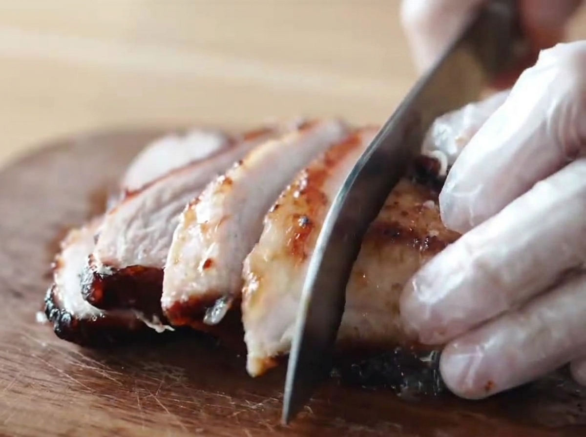 Ein karamellisiertes, honigglasiertes Stück geräuchertes Chashu-Schweinefleisch wird von einer Person mit Handschuhen auf einem Holzbrett in dicke Scheiben geschnitten.