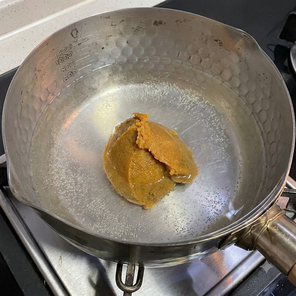 Ein Löffel weiße Misopaste löst sich in einem Topf mit heißem Wasser auf einem Herd auf.