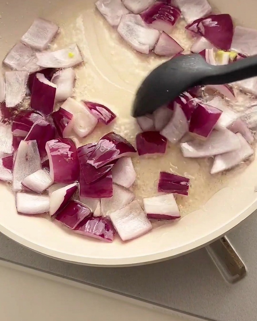 Gewürfelte rote Zwiebeln werden mit einem schwarzen Spatel in einer geölten Pfanne angebraten.