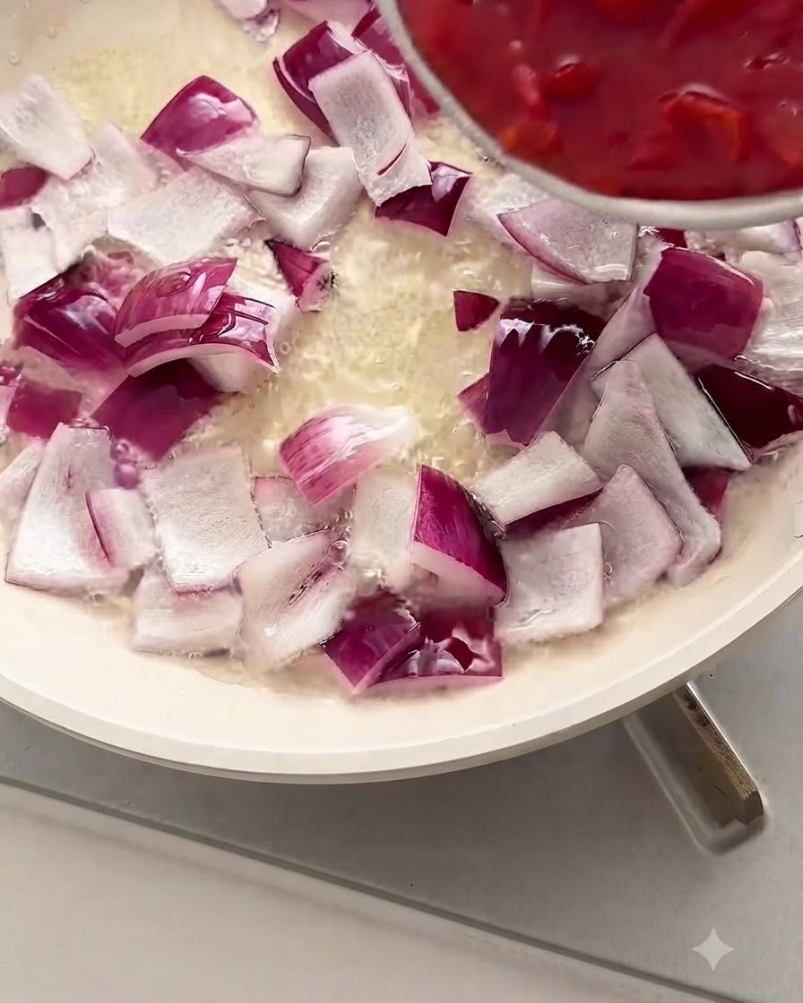 Gehackte rote Tomaten werden aus einer Schüssel in eine weiße Pfanne mit gewürfelten roten Zwiebeln und Öl gegeben.