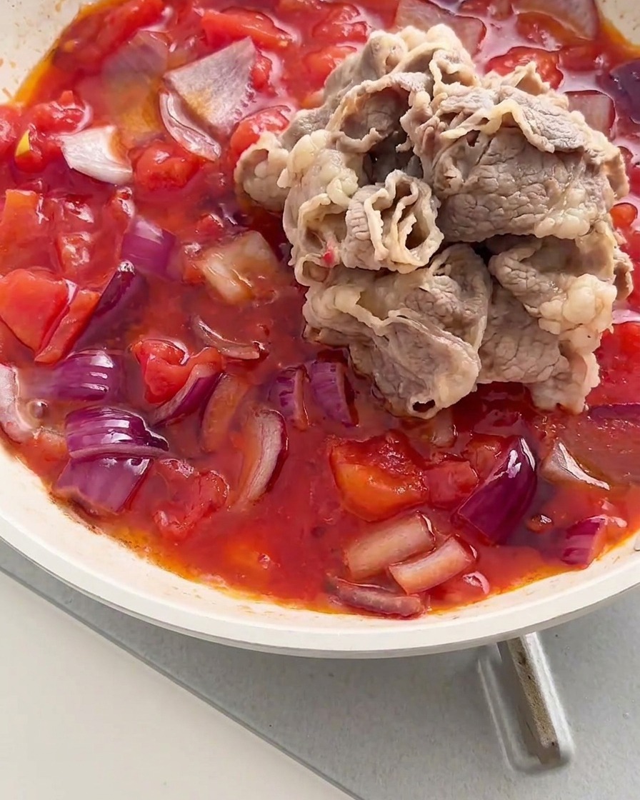 Ein Haufen blanchierter Rindfleischröllchen wird in eine Pfanne mit einer köchelnden Mischung aus roten Tomaten und Zwiebeln gegeben.