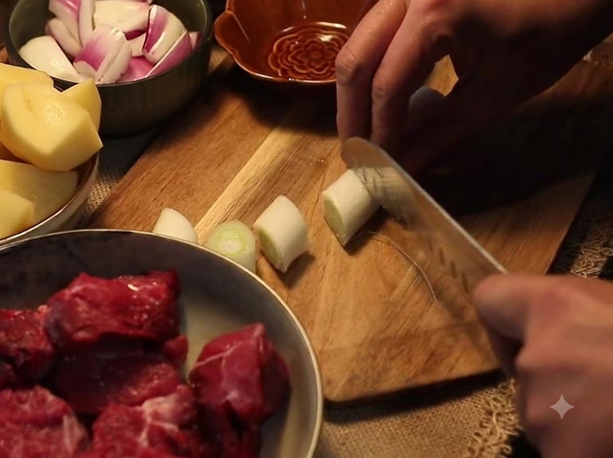 Ein Messer schneidet eine Lauchzwiebel in dicke Abschnitte auf einem Holzschneidebrett neben Schüsseln mit vorbereitetem Gemüse und rohem Rindfleisch.