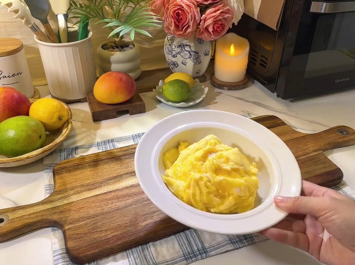 Ein Teller mit weichem Rührei auf einem Holzbrett neben frischen Früchten und Küchenutensilien.
