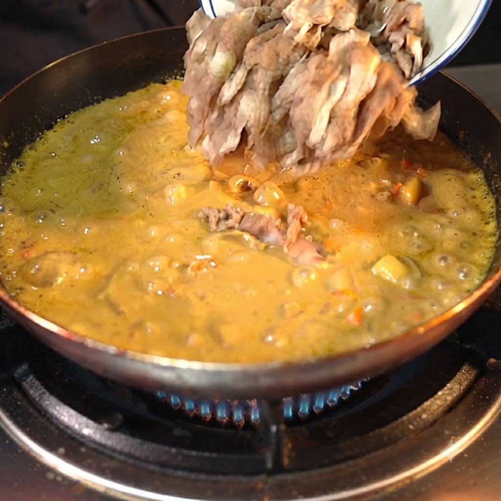 Vorbereitete Rindfleischscheiben werden aus einer weißen Schale in eine köchelnde, eingedickte gelbe Currysauce in einer dunklen Pfanne gegossen.